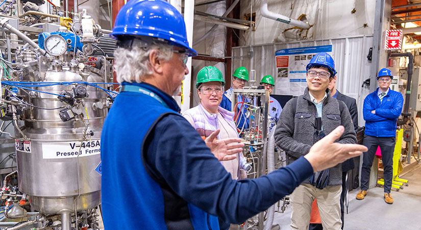 Six people wearing hard hats and safety glasses on a tour in a lab.